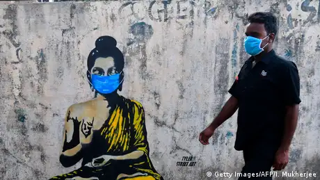 A graffiti of the Buddha wearing a blue facemask with a person walking next to him wearing the same facemask.