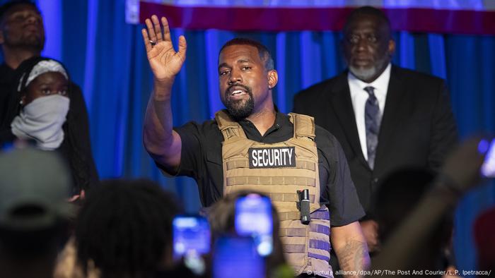 Wearing a security vest, Kanye West holds up his hand and addresses an audience 