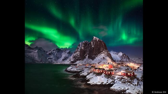 Night shot of a small coastal settlement with red houses and snow capped mountains.  In the sky you can see sparkling green lights (Photo: Andreas Ettl).