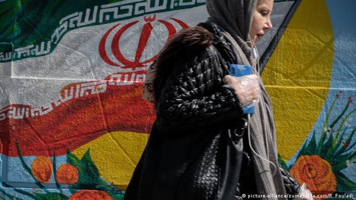 Iranians wearing face masks and single-use gloves walk past a mural displaying their national flag in Tehran
