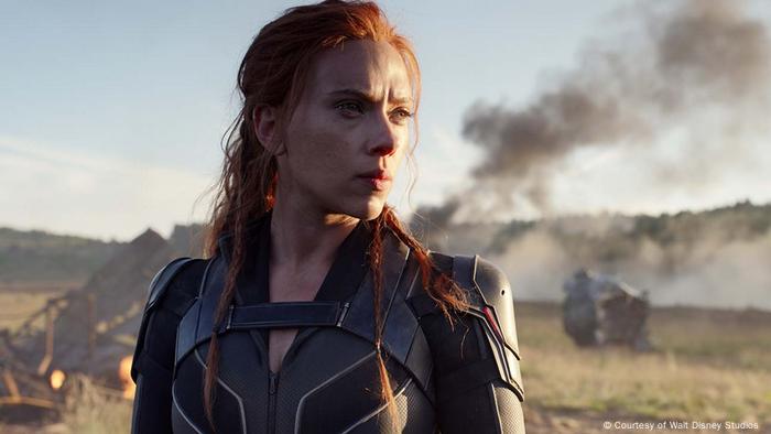 Scarlett Johansson as the superheroine the Black Widow, stands in a smoking wasteland, wearing a futuristic costume and looking pensive.