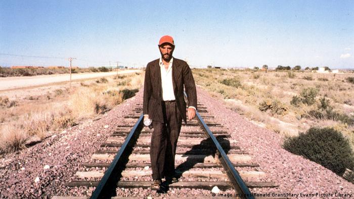 Filmszene aus Paris, Texas von Wim Wenders: Ein Mann mit orangefarbener Kappe und Anzug läuft in der Wüste an einem Bahngleis entlang (Bild: imago images/Ronald Grant/Mary Evans Picture Library)