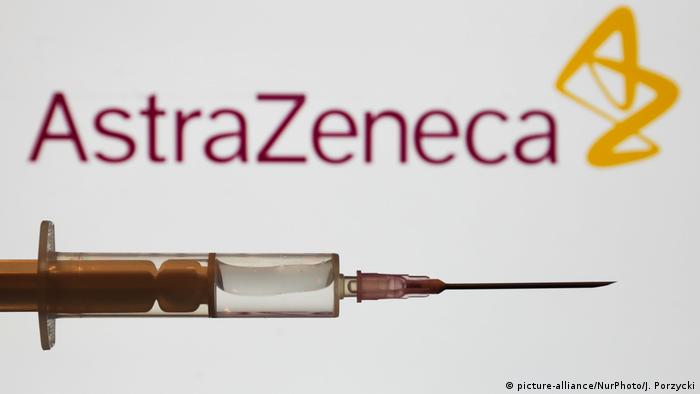 A medical syringe is seen with the AstraZeneca company logo displayed on a screen in the background