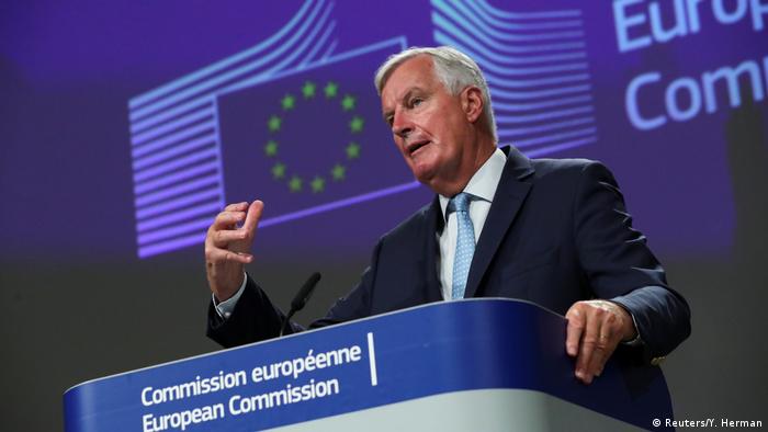Michel Barnier stands at a podium wearing a dark blue suit and with his right hand slightly raised