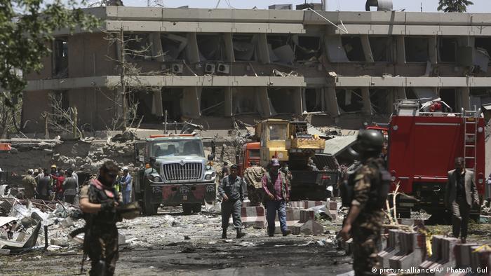 Blick auf ein langes, graues Gebäude mit Zerstörungen durch eine Bombenanschlag, davor Trümmerteile, LKWs, Arbeiter und Soldaten, die das Gelände sichern