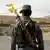 Blick auf den Rücken eines dunkelhaarigen Manns in Uniform an einer Straße mit einem Gewehr, aus dessen Lauf ein Zweig mit gelben Blumen ragt. Im Hintergrund kahle Hügel und einige weiße Flachbauten