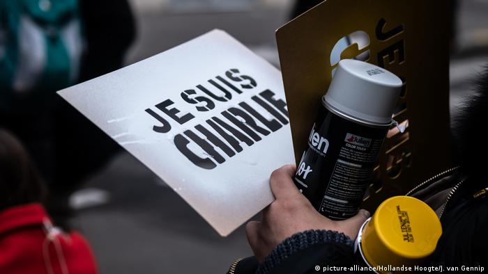 Hands hold a placard that reads Je suis Charlie, and a spray can
