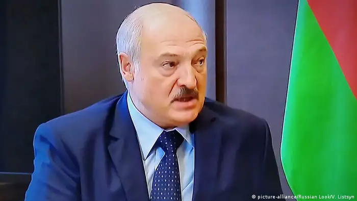 A close up of Lukashenko's face next to a Belarus flag. He has a small mustache and is wearing a blue jacket, light blue shirt and a spotted blue tie. 