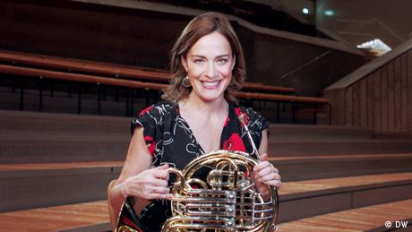 Sarah Willis holds a french horn, smiling. She is sitting in a rehearsal hall. 