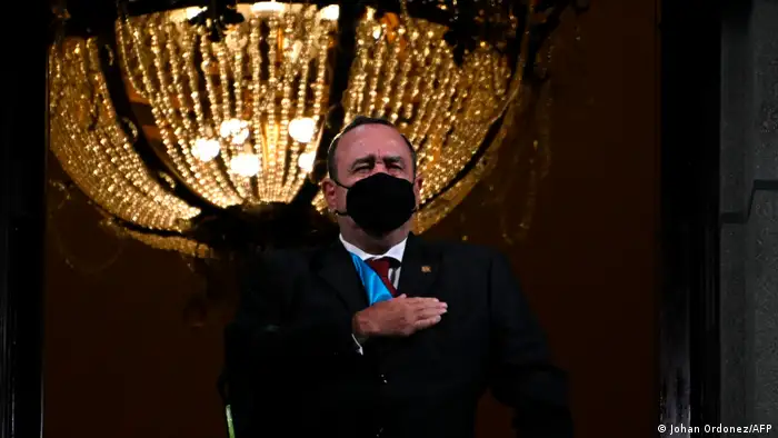 Alejandro Giammattei wears a black face mask and stands in front of a large chandelier