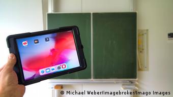 Eine Hand hält in einem leeren Klassenzimmer ein Tablet vor eine Schultafel.