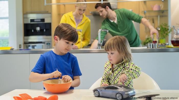 Little girl and boy at a table while mother and father cook in the kitchen behind them