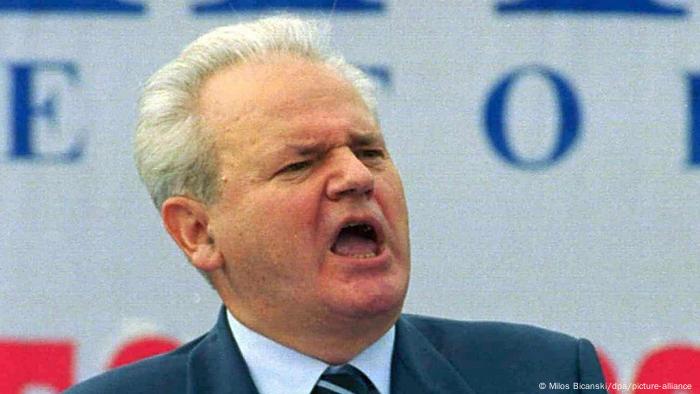Ein Mann mit grauen Haar spricht oder schreit mit offenem Mund, hinter ihm sind blaue und weiße Buchstaben auf weißem Grund zu sehen