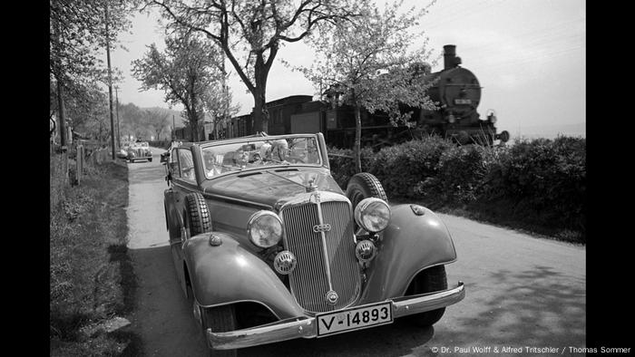 Auto steht auf einer Straße während im Hintergrund ein Zug anrauscht (Dr. Paul Wolff & Alfred Tritschler / Thomas Sommer)