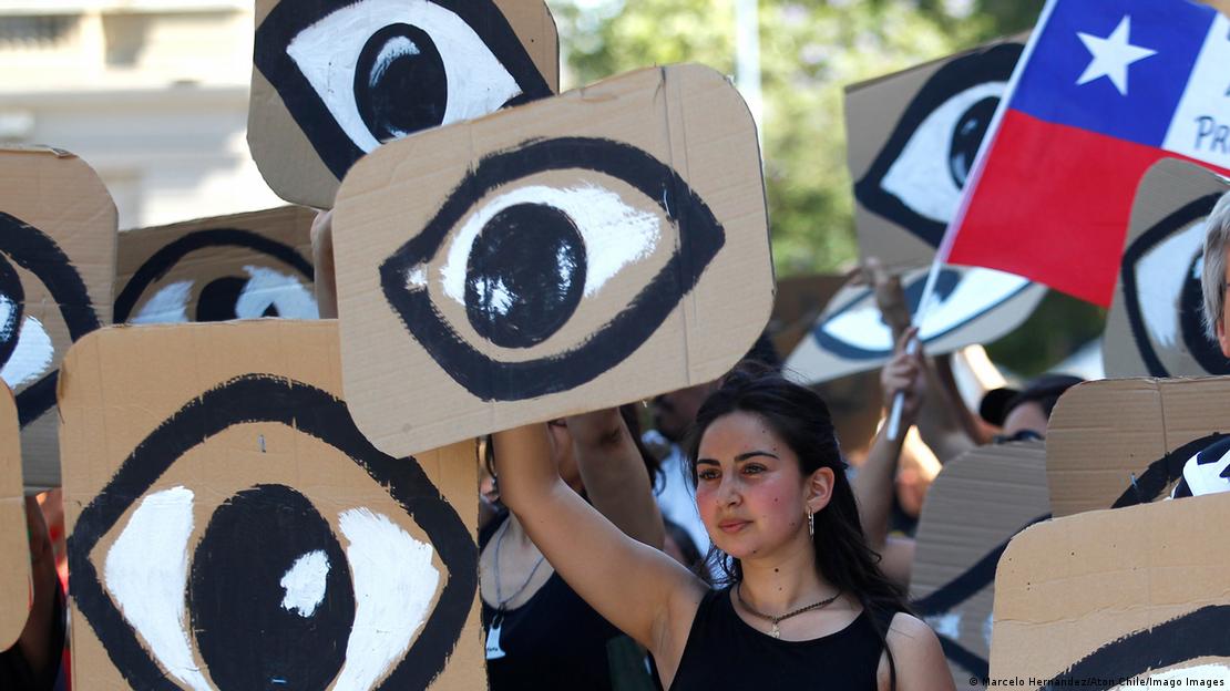 Jóvenes manifestantes portan 352 ojos de cartón, simbolizando a quienes perdieron los suyos en la represión del estallido social en Chile. Entre los ojos, una bandera chilena. Jóvenes manifestantes portan 352 ojos de cartón, simbolizando a quienes perdieron los suyos en la represión del estallido social en Chile. Entre los ojos, una bandera chilena.