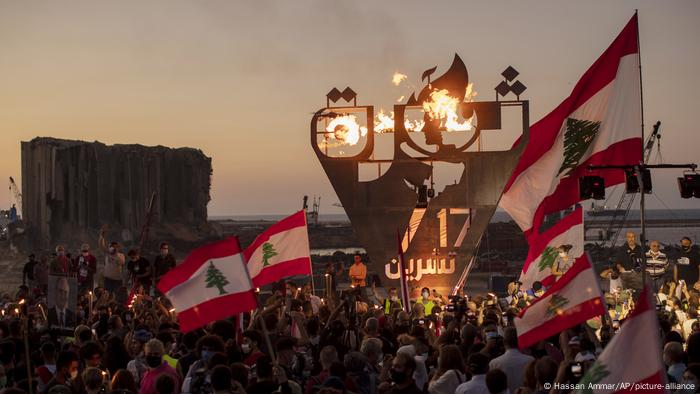 Protesters wave Lebanese flags in the twilight in front of a 'October 17 revolution' sign and the remains of Beirut's destroyed port