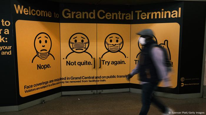 Homem de máscara caminha em frente a grande cartaz que explica como usar a máscara corretamente. 