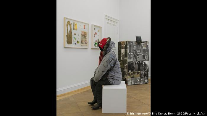 A human-sized sculpture sits on a bench in an art gallery facing a wall covered with paintings (Iris Kettner/VG Bild-Kunst, Bonn, 2020/Foto: Nick Ash)