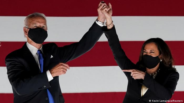 Joe Biden and Kamala Harris hold hands in front of a giant American flag while wearinmasks