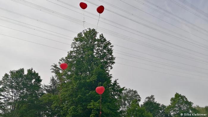 Vor der Kulisse grüner Laubbäume und einem blaugrauen Himmel steigen vier rote Luftballons in Herzform nach oben. Tod und Trauer in der Corona-Zeit