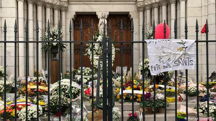 Muitas flores em frente à igreja. Há um portão, que está fechado. No portão, está pendurado com cartaz com uma frase em francês e o desenho de uma pomba. 