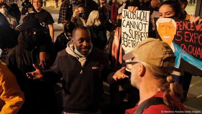 An African American man holds out his hands while in discussion with a white man with a hat on, outdoors