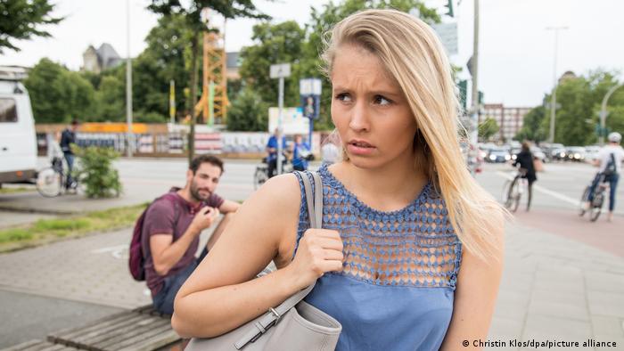 A young woman on a street looks upset as a man behind her watches her