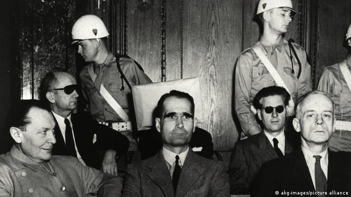 Nürnberger Prozess: Die Angeklagten Göring, Heß, von Ribbentrop, Dönitz, von Schirach sitzen sitzen in zwei Reihen, hinter ihnen stehen Wächter (akg-images/picture-alliance).