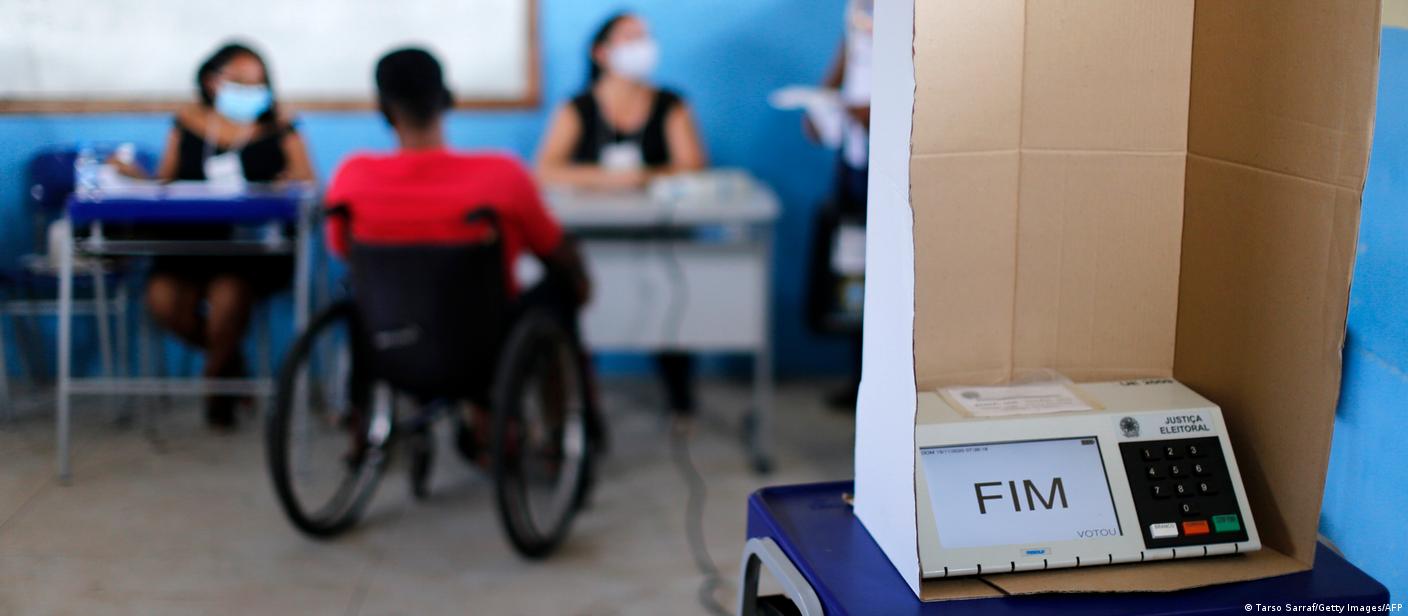 Urna eletrônica com cadeirante ao fundo, diante de mesários em local de votação