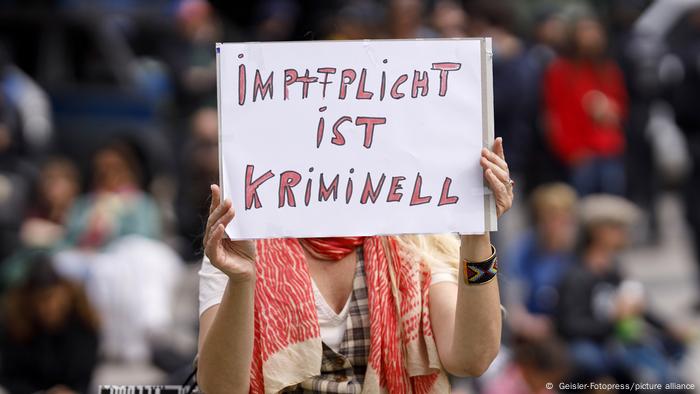 Woman in a crowd, holding a handwritten sign in front of her face, which reads mandatory vaccination is a crime (in German)