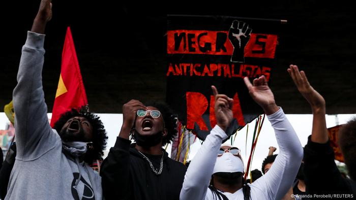 Pessoas negras gritam e gesticulam diante de faixa Negros anticapitalistas Pessoas negras gritam e gesticulam diante de faixa Negros anticapitalistas