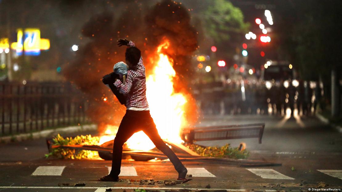 Homem no meio da rua em frente a fogueira, durante protesto noturno Homem no meio da rua em frente a fogueira, durante protesto noturno