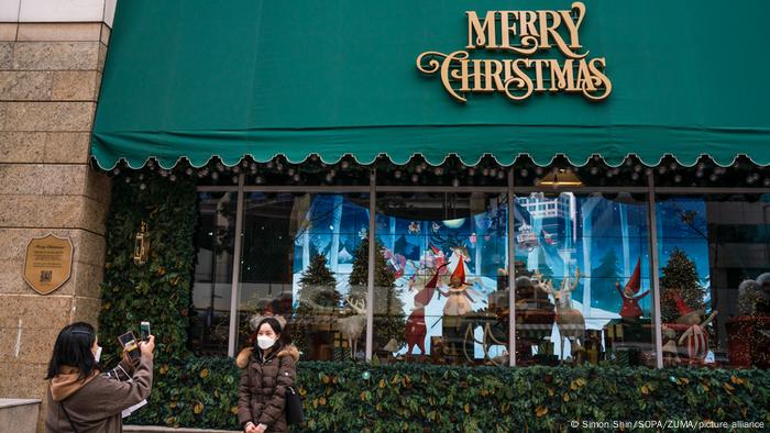 Asiatische Frau mit Maske steht vor einer weihnachtlichen Schaufenster-Dekoration, eine weitere Frau fotografiert sie.