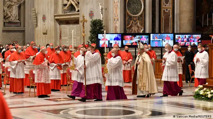 Der Papst und mehrere Würdenträger in weißen Gewändern schreiten an Männern in scharlachroten Talaren vorbei 