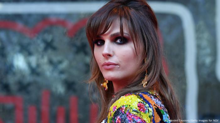 Andreja Pejic looking over her shoulder at the camera. She has long brown hair, dark eye-shadow and wears a colorful top.