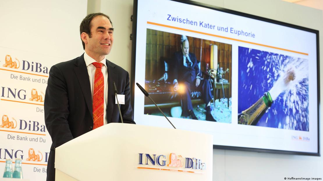 Carsten Brzeski me kostum të zi dhe kravatë të kuqe duke folur në pult