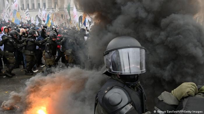 A police officer in riot gear and a helmet stands in a cloud of black smoke