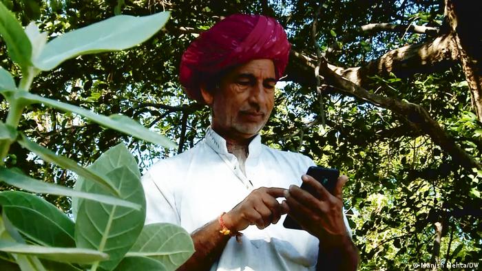 A man stands in the shadows underneath a tree and types into his mobile phone in India