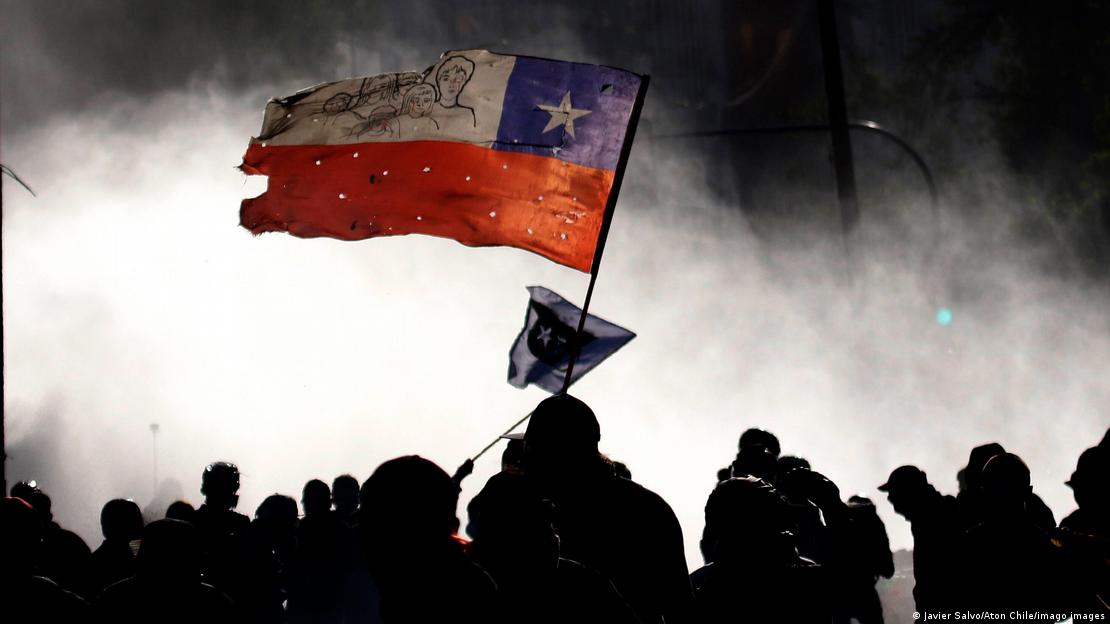 Manifestantes en medio del gas lacrimógeno lanzado por la policía. Sobre ellos, una bandera de Chile.