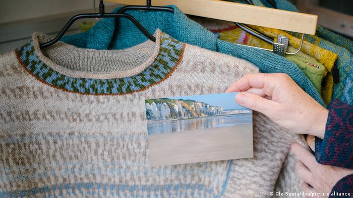 A hand holds up a photo of Normandy coast against a knit sweater that uses the same color scheme