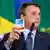 Bolsonaro, de terno e gravata, segura uma caixa de hidroxicloroquina. Ao fundo, desfocada, a bandeira do Brasil. 
