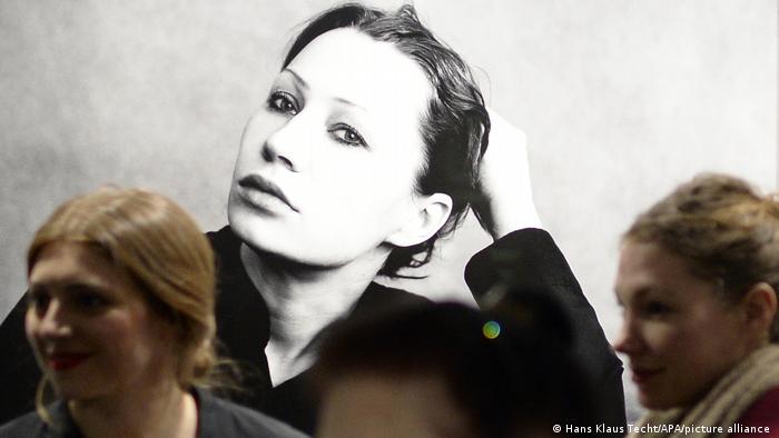 Schwarz-Weiß Porträt von Schauspielerin Birgit Minichmayr: Sie stütz ihren Kopf auf ihrem Arm ab und schaut in die Kamera. Davor Musesumsbesucherinnen