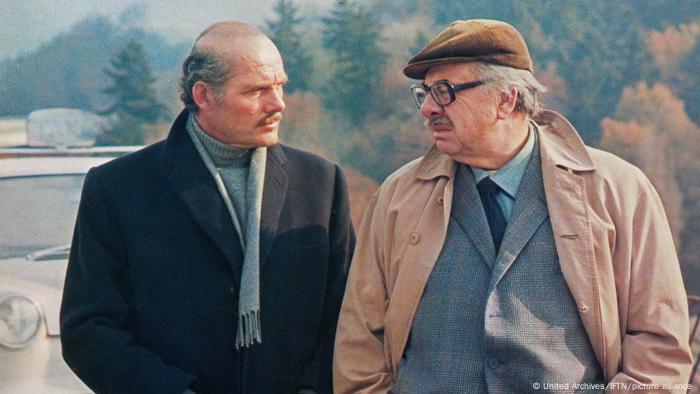 Two elderly men in suits and jackets face each other, trees in the background