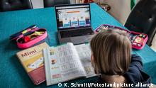 Bamberg, Deutschland 04. Januar 2021: Ein Kind sitzt an einem Tisch in einer Wohnung auf dem ein aufgeklappter Laptop mit einem Online-Lernprogramm steht. Neben dem Laptop liegen verschiedene Schulutensilien für das Homeschooling. Das Kind löst Aufgaben in einem Schulheft.