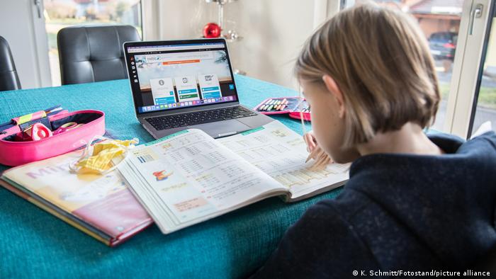 Auf einem Wohnzimmertisch steht ein Laptop an dem gerade ein Kind arbeitet, daneben liegen verschiedene Schulutensilien, ein aufgeschlagenes Buch und ein Mundschutz.