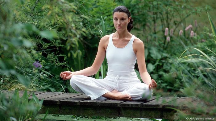 Woman wearing white clothes sits, legs crossed, on a wood bridge surrounded by plants