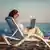 سيدة تجلس على شاطئ وبين يديها جهاز كمبيوتر لوحي (صورة رمزية)