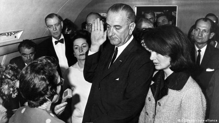 Der US-amerikanische Vizepräsident Lyndon B. Johnson steht mit erhobener Hand neben Jacqueline Kennedy, umringt von mehreren Leuten 
