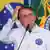 Bolsonaro, de roupa branca, coça a cabeça. Ele está atrás de um microfone. Atras do presidnete, é possível ver a bandeira do Brasil. 