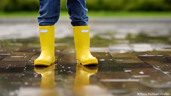 Legs of child wearing yellow rubber boots standing on pavement covered in water